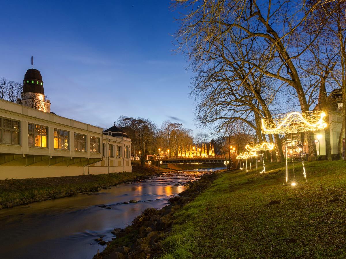 Beleuchtetes Gradierwerk und Bega in Bad Salzuflen bei Dämmerung mit festlichen Lichtern am Ufer und auf der Brücke.