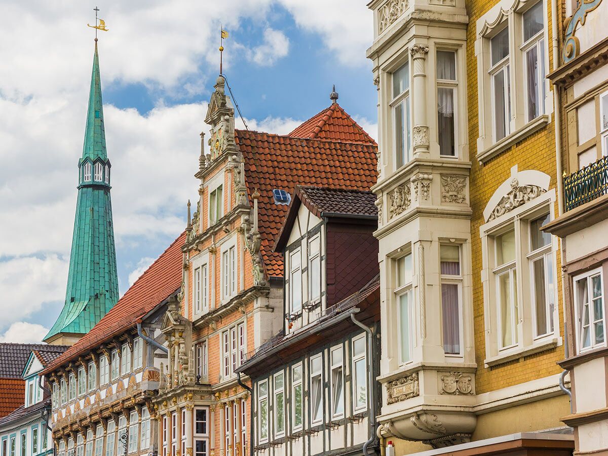 Detailreiche historische Gebäude und ein grüner Kirchturm mit Wetterfahne in einer deutschen Altstadt unter blauem Himmel.