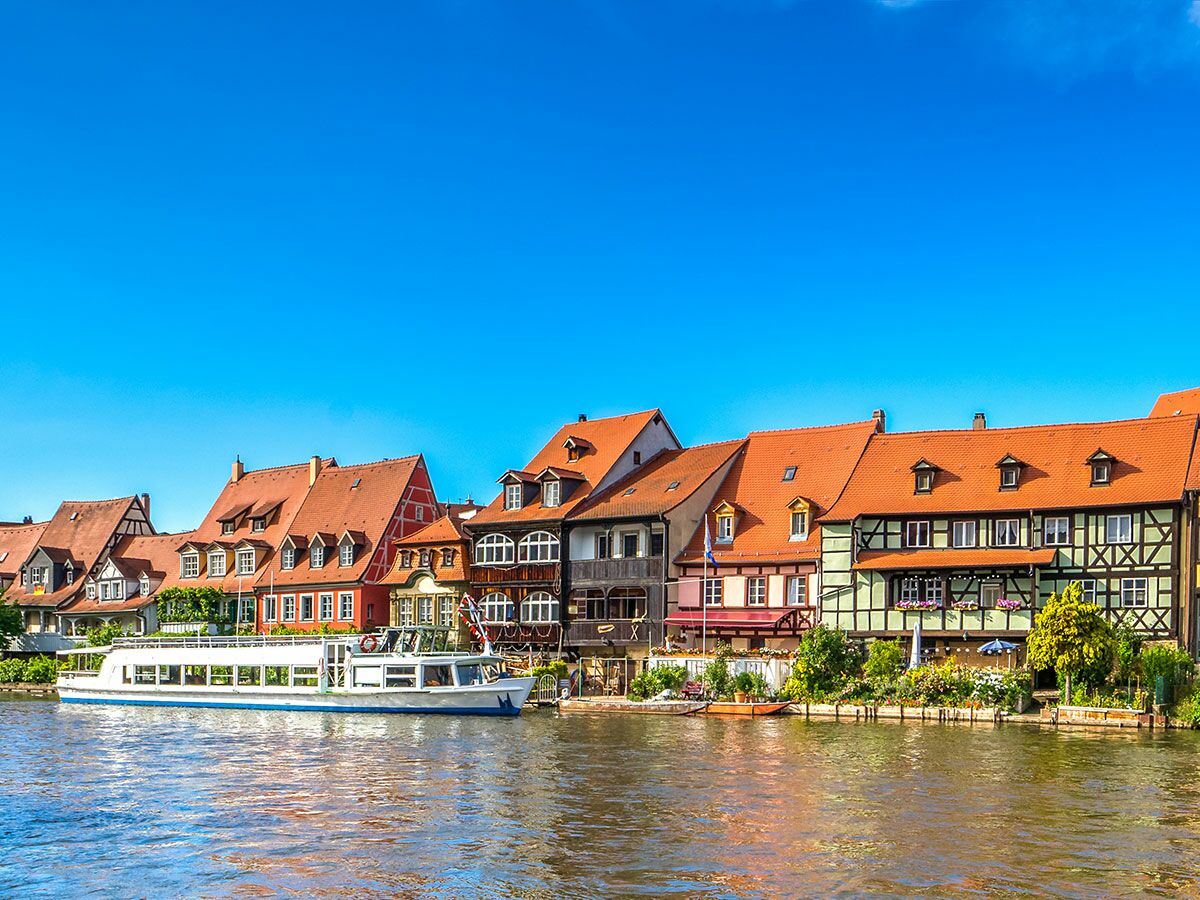 Idyllisches Flussufer in Bamberg mit Fachwerkhäusern, roten Dächern und einem großen Ausflugsboot auf dem Wasser.