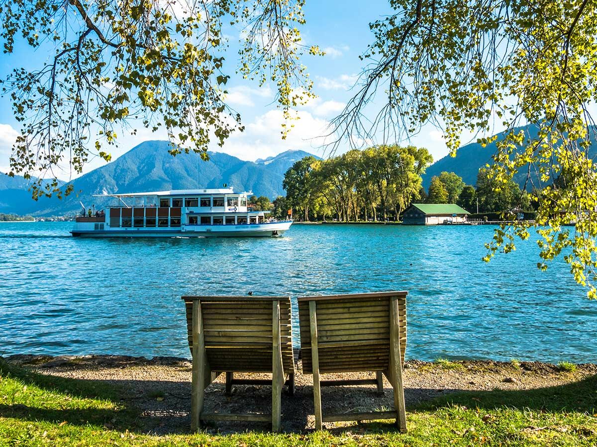 Blick über einen ruhigen Bergsee mit Passagierschiff, umrahmt von herbstlichen Bäumen und Sitzbänken am Ufer.
