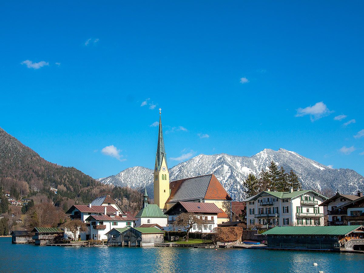 Malerisches Rottach-Egern am Tegernsee mit St. Laurentius Kirche, Bootshäusern, schneebedeckten Alpen und blauem Himmel im Frühling.