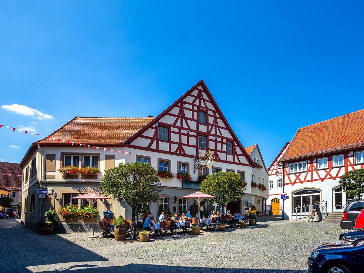 Malerischer deutscher Marktplatz mit Fachwerkhäusern, Außengastronomie, Blumenkästen, blauer Himmel und gepflastertem Boden.