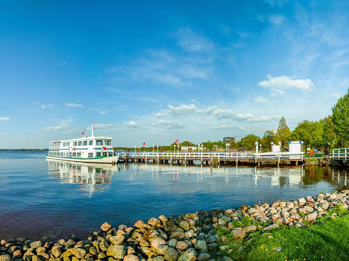 Weißes Passagierschiff am hölzernen Anleger bei sonnigem Wetter an einem See, mit blauem Himmel und Felsen am Ufer.