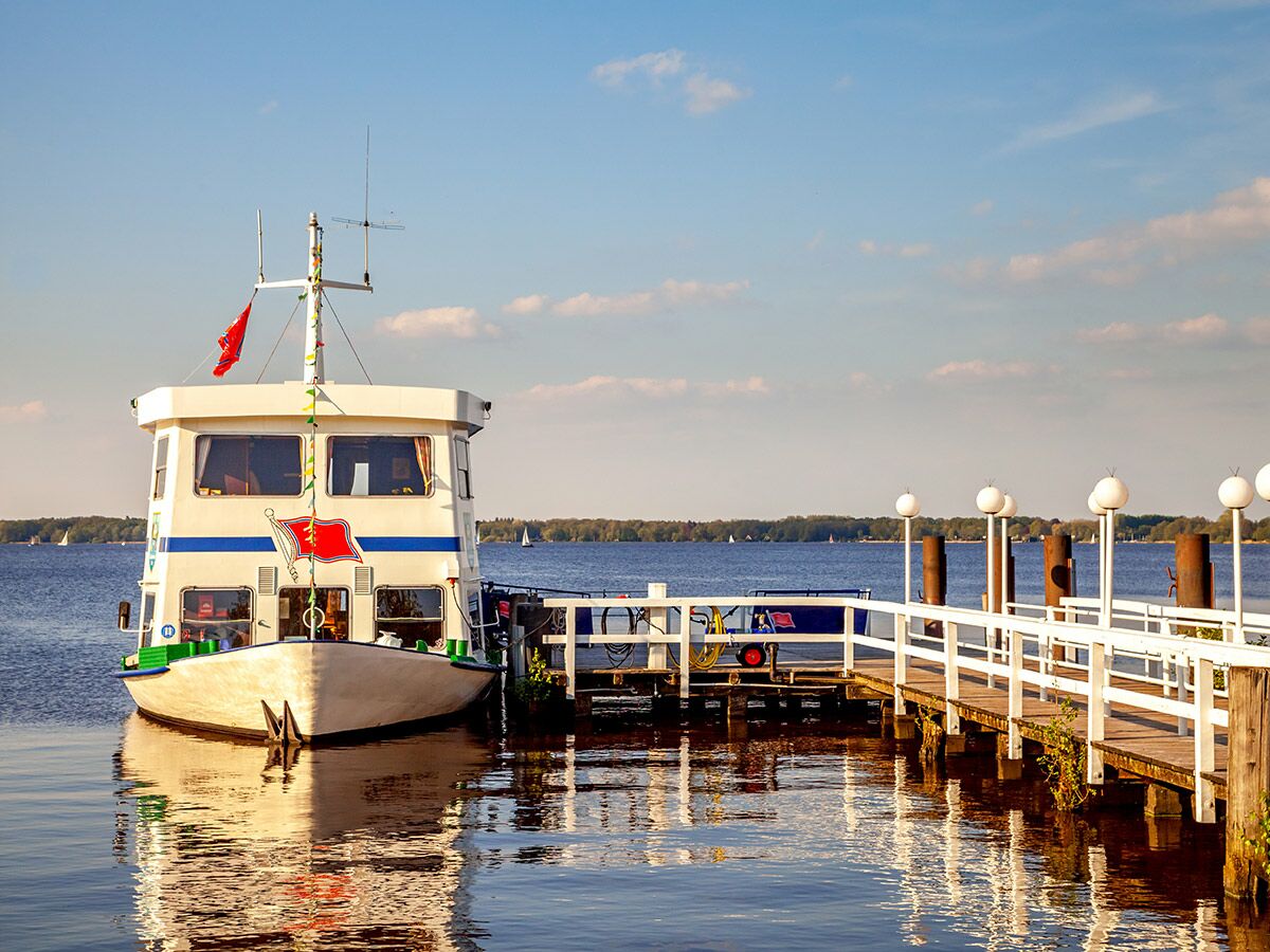 Ein weißes Passagierboot liegt an einem Holzsteg auf einem ruhigen See unter blauem Himmel mit leichten Wolken.