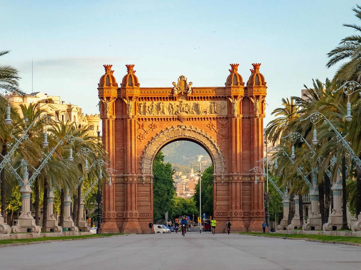 Der Arc de Triomf in Barcelona im Morgenlicht, umgeben von Palmen und Menschen, die Fahrrad fahren und spazieren gehen.