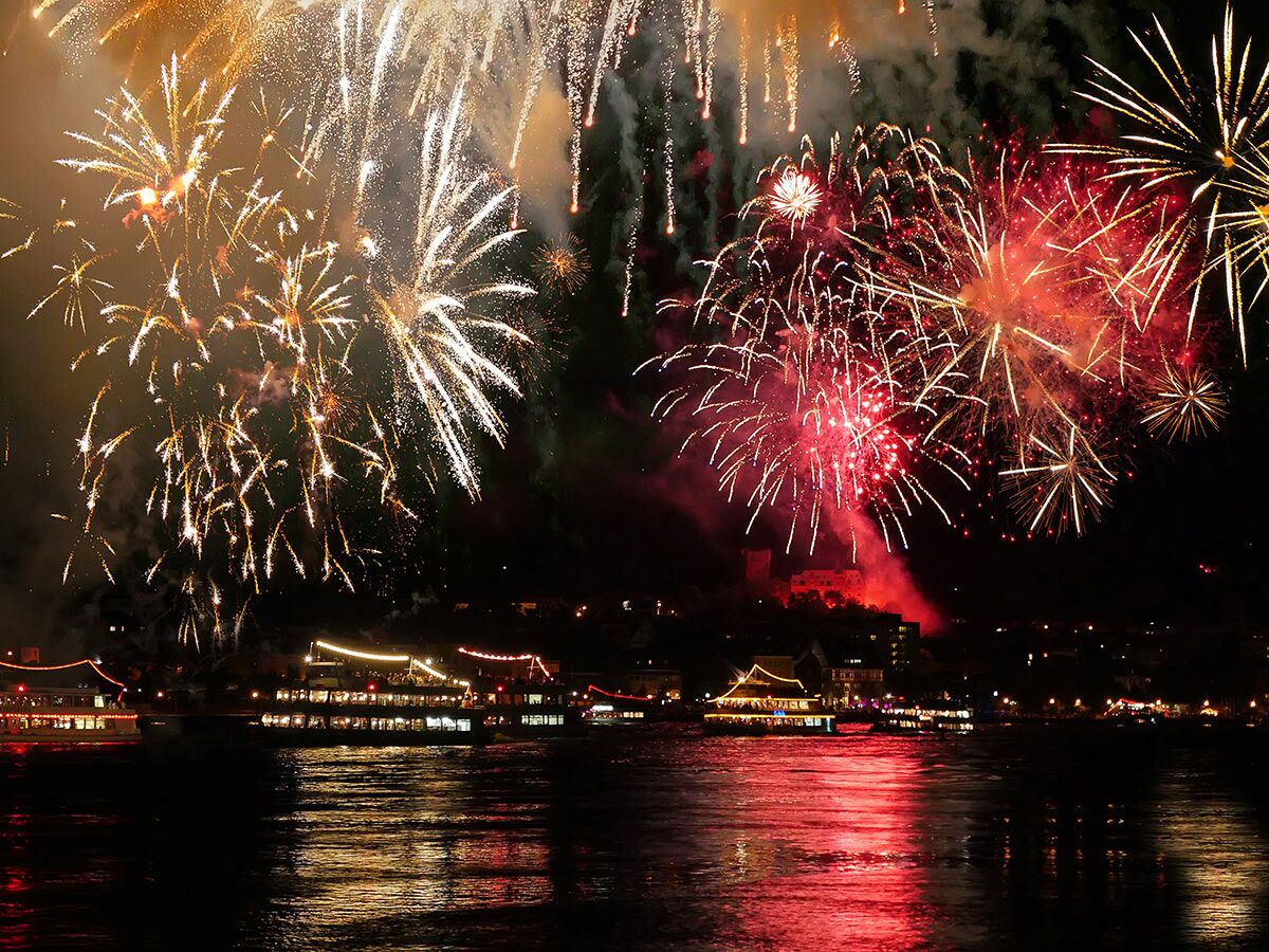 Spektakuläres Feuerwerk in Rot und Gold über einer beleuchteten Stadt am Flussufer mit historischen Gebäuden und Booten.
