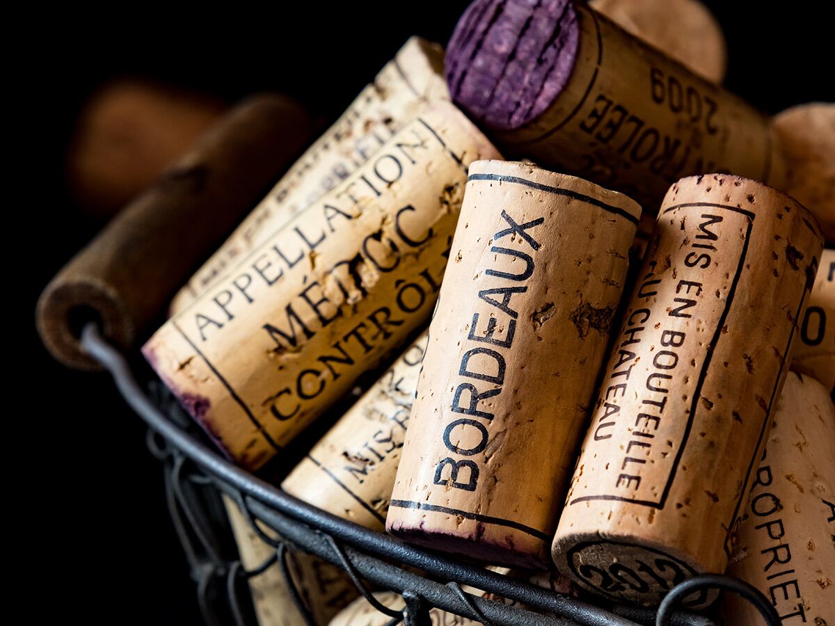 Nahaufnahme von vielen Weinkorken mit Aufschriften wie "Bordeaux" und "Appellation Médoc Contrôlée" in einem Drahtkorb.