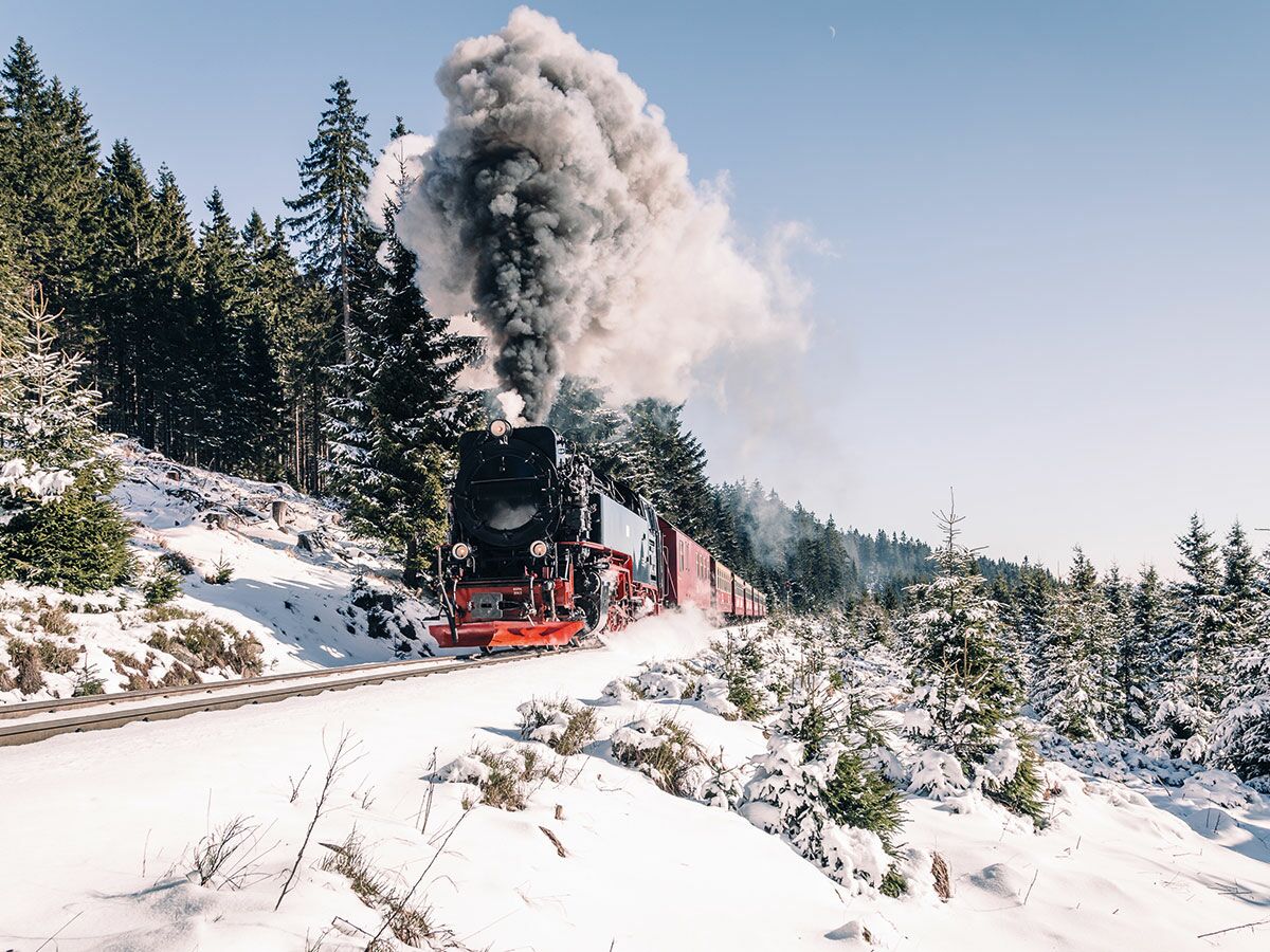 Schwarzer Dampfzug mit roten Akzenten fährt durch tief verschneite Winterlandschaft mit Nadelbäumen unter blauem Himmel, stark rauchend.