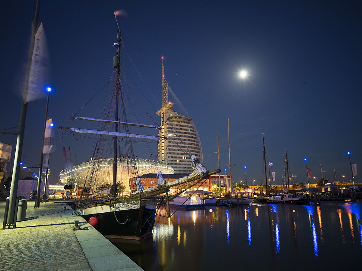 Nachtaufnahme des Hafens in Bremerhaven mit großem Segelschiff, modernem Gebäude, Mond und leuchtenden Wasserreflexionen.