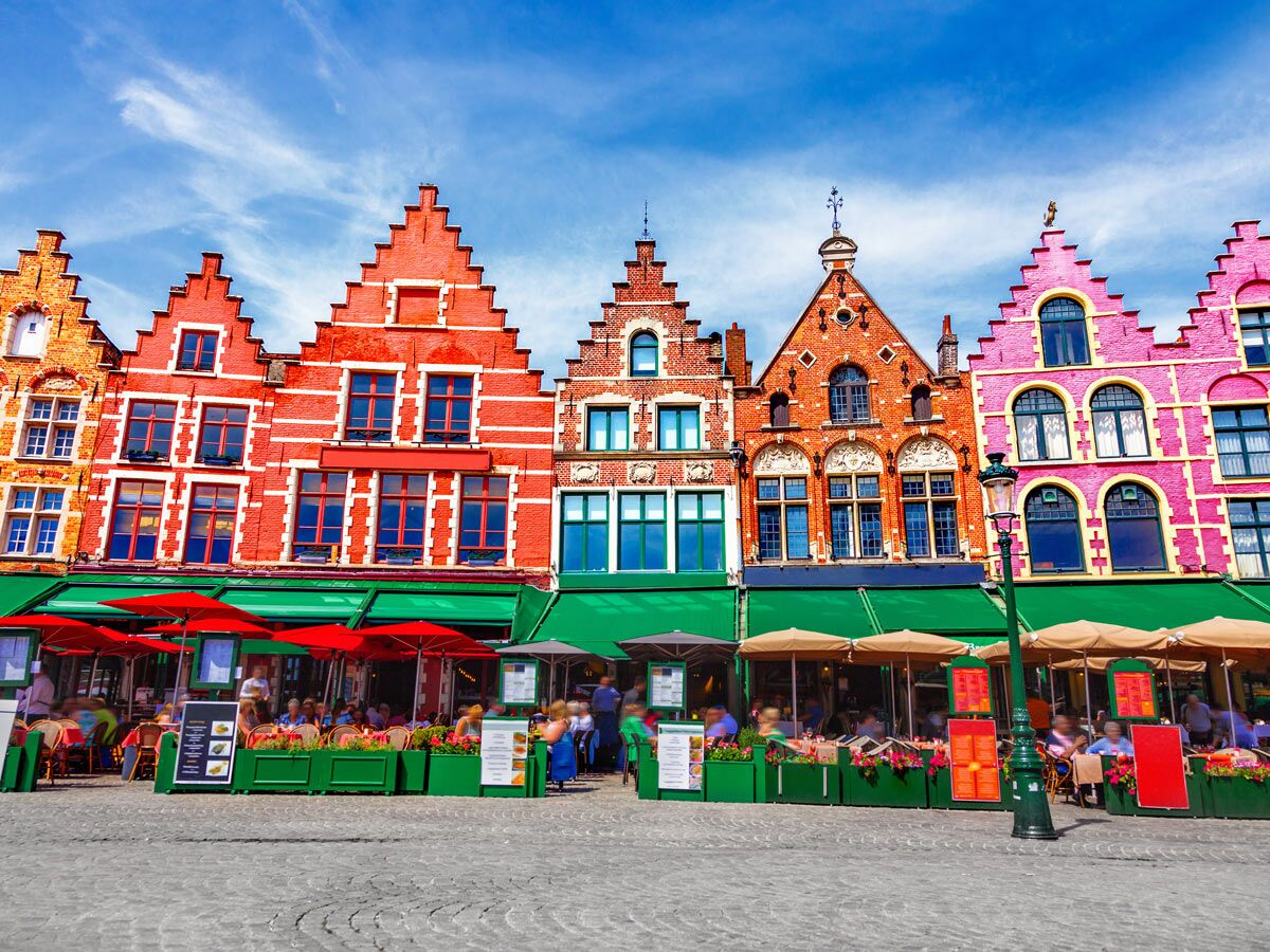 Bunte Giebelhäuser am Marktplatz von Brügge mit lebhaften Straßencafés, grünen Markisen und roten Sonnenschirmen unter blauem Himmel.