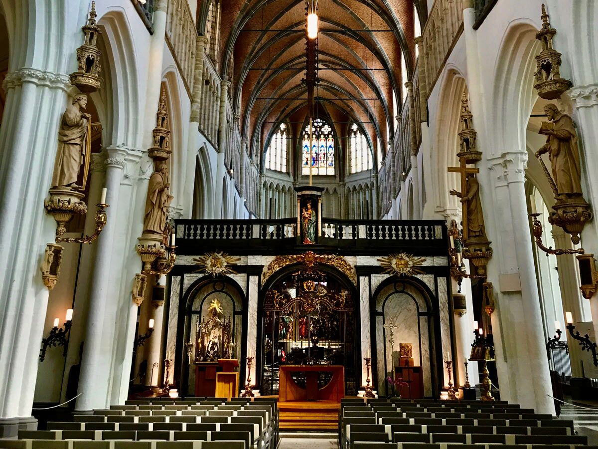 Innenansicht einer gotischen Kathedrale mit hohen Gewölbedecken, weißen Säulen, Statuen, goldenem Altar und Sitzreihen.