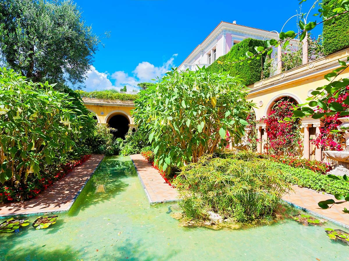 Wunderschöner mediterraner Garten mit grünem Teich, üppigen Pflanzen, bunten Blumen und eleganter Villa unter blauem Himmel.