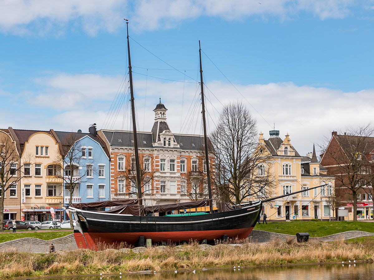 Buntes Hafenviertel in Emden mit dem historischen Schiff Greundiek am Kanal und detailreichen Gebäuden unter klarem Himmel.