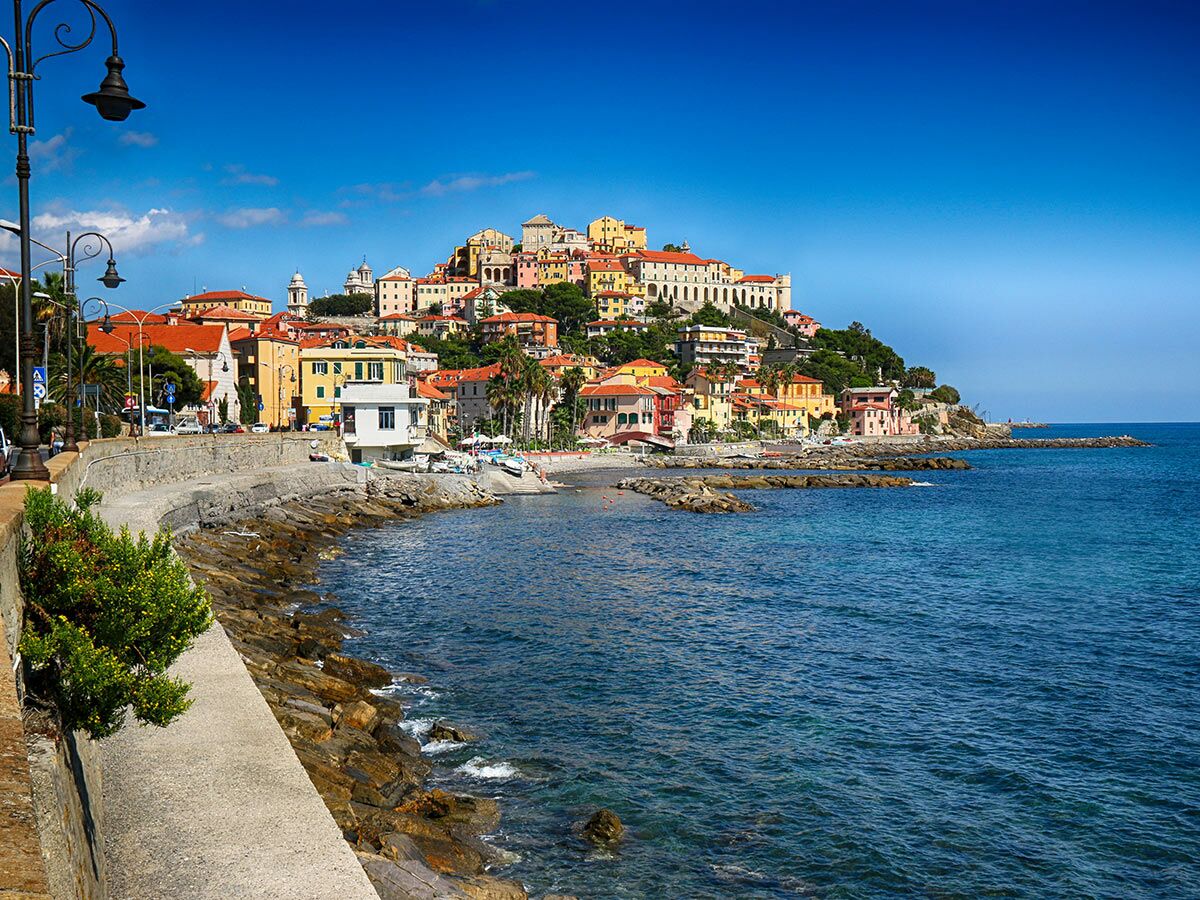 Panoramablick auf eine italienische Küstenstadt mit bunten Häusern am Hang, tiefblauem Mittelmeer und felsiger Küste unter strahlend blauem Himmel.