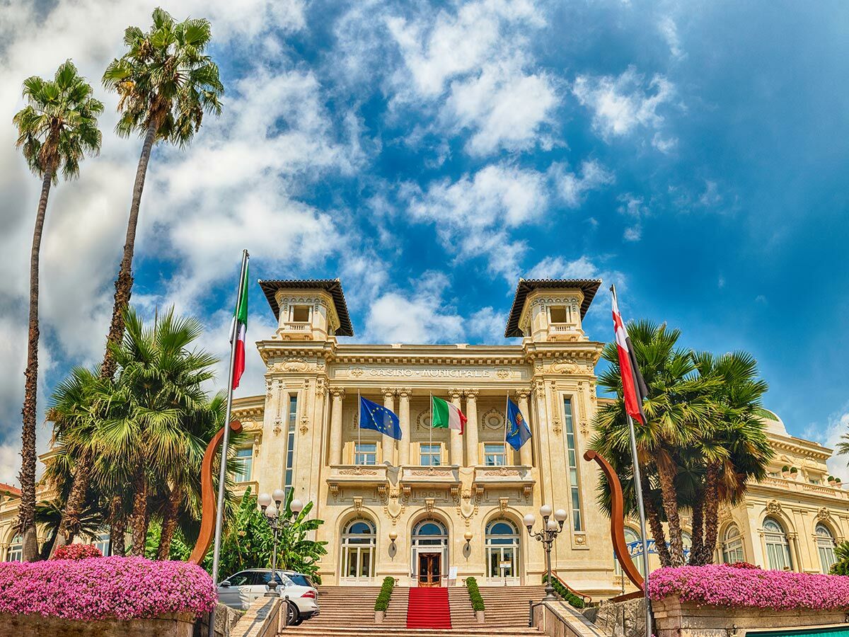 Historisches Casino Sanremo mit Palmen, italienischer Flagge und blauem Himmel. Elegant, repräsentativ, sonnig.