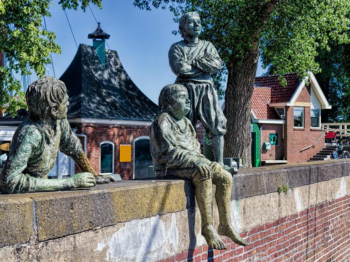 Drei Bronzestatuen von Jungen, einer lehnend, einer sitzend, einer stehend, an einer Steinmauer vor Backsteingebäuden.