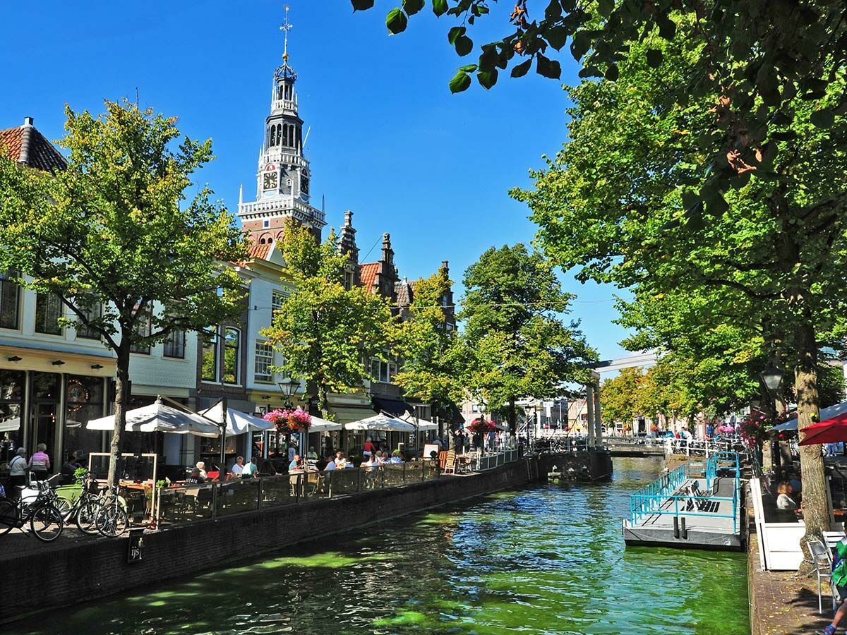 Historische Stadtansicht Gouda mit Gracht, belebten Terrassencafés, grünen Bäumen und dem Rathaus-Glockenturm bei Sonnenschein.