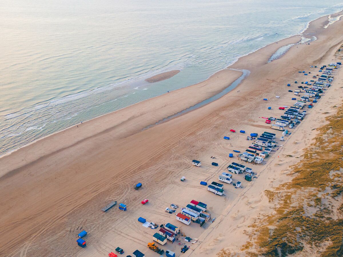 Luftaufnahme eines weiten Sandstrandes mit einer langen Reihe von Wohnwagen und Strandhütten am Meer und Dünen.
