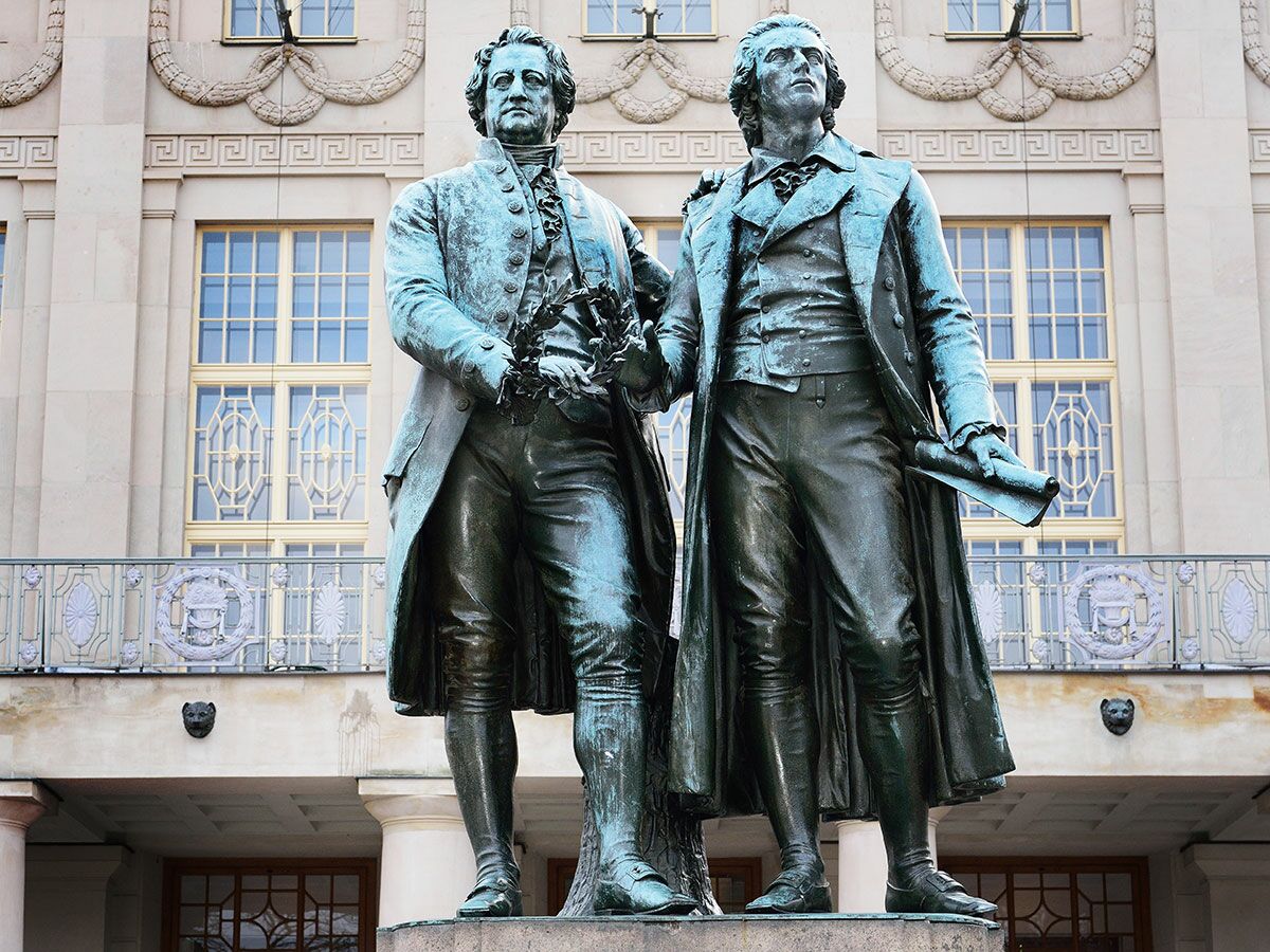 Bronze-Statuen von Goethe und Schiller vor einem historischen Gebäude. Goethe hält Lorbeerkranz, Schiller eine Schriftrolle.