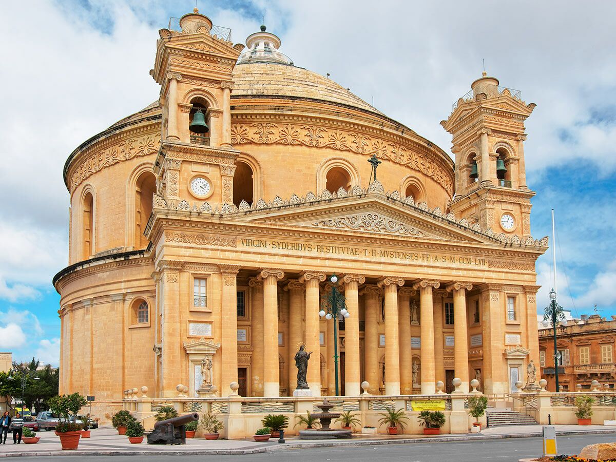 Die Rotunde von Mosta, eine beeindruckende beige-farbene Kuppelkirche in Malta, mit Säulenfassade, zwei Glockentürmen und blauem Himmel.