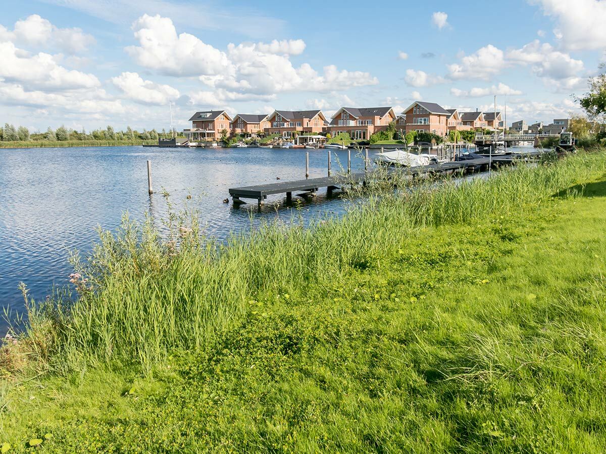 Moderne Backsteinhäuser an einem See mit Bootsstegen, umgeben von grüner Natur unter blauem Himmel.