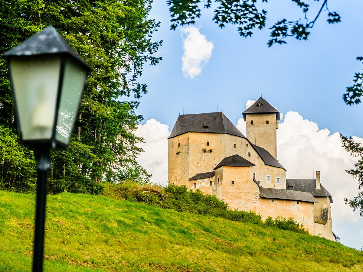 Historische Burg mit Türmen auf einem bewaldeten Hügel unter blauem Himmel mit Wolken, im Vordergrund Laterne und Gras.