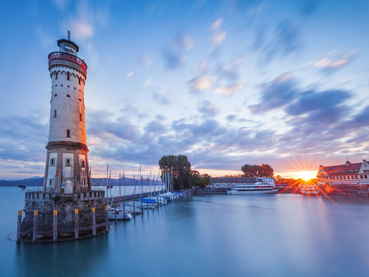 Der Lindauer Leuchtturm und der Hafen am Bodensee bei Sonnenaufgang mit Booten und Bergen im Hintergrund.