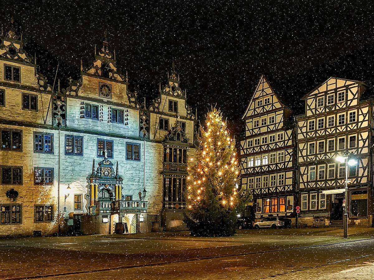 Verschneiter Weihnachtsmarkt in einer historischen Altstadt bei Nacht mit beleuchtetem Weihnachtsbaum und Fachwerkhäusern.
