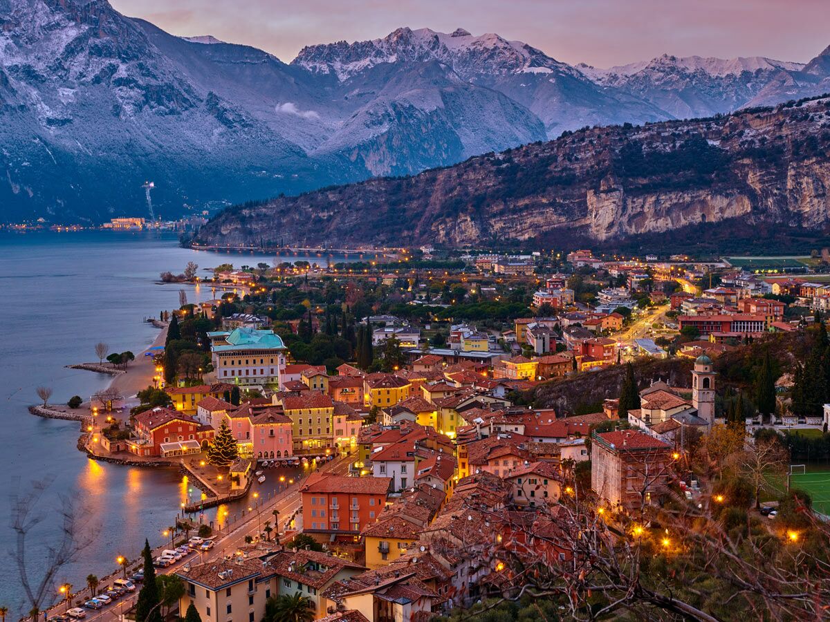 Abendlicher Blick auf eine beleuchtete Stadt am Gardasee mit schneebedeckten Bergen und festlicher Weihnachtsbeleuchtung.