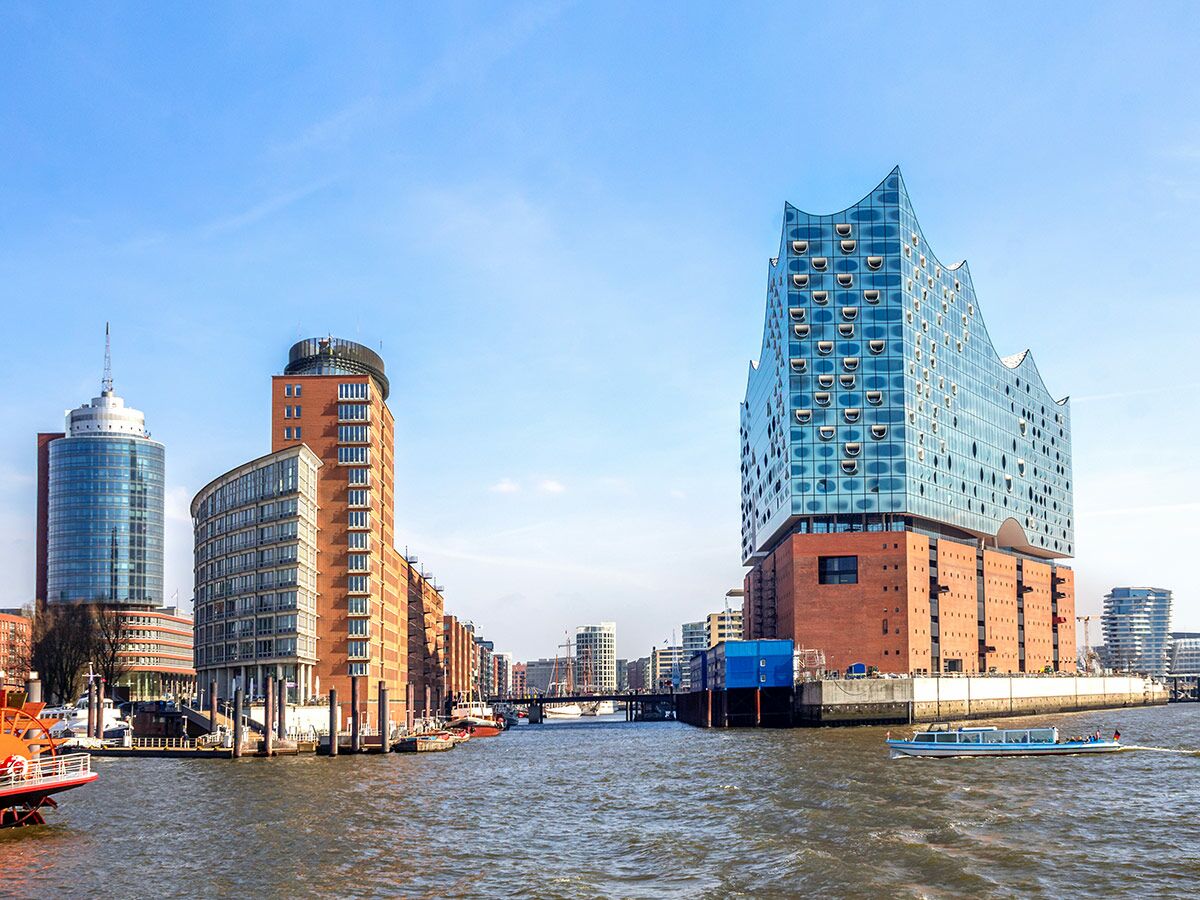 Panoramablick auf die Hamburger Elbphilharmonie mit weiteren Gebäuden, Booten und blauem Himmel über dem Wasser.