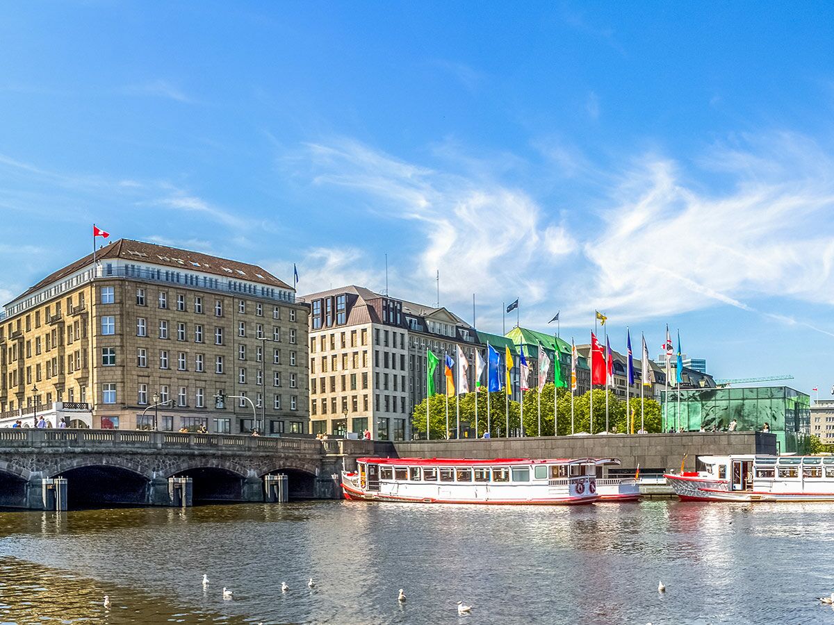 Boote auf Fluss, historische Brücke und Gebäude in Hamburg. Internationale Flaggen wehen unter klarem blauem Himmel.