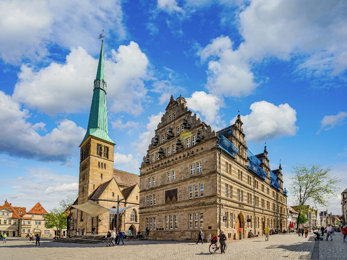Historischer Marktplatz mit hoher Kirche und Rathausgebäude, Menschen flanieren unter blauem Himmel mit weißen Wolken.