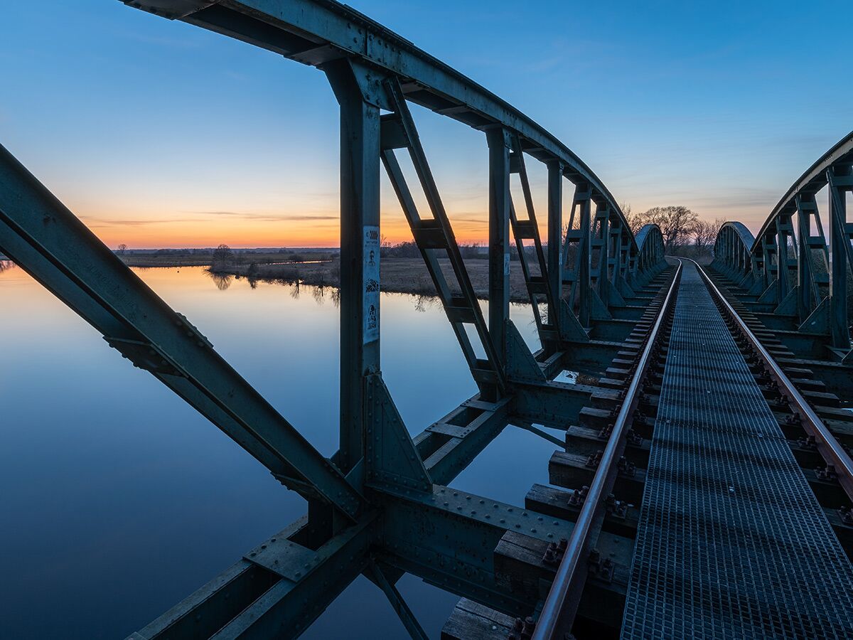 Weitwinkelaufnahme einer Stahl-Eisenbahnbrücke über einen Fluss bei Sonnenuntergang mit spiegelndem Wasser und goldenem Himmel.