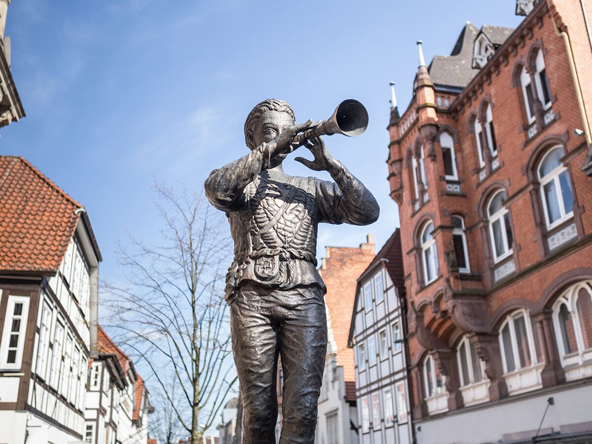 Bronze Rattenfänger Statue bläst Horn in Hameln vor historischen Fachwerkhäusern und rotem Backsteingebäude unter blauem Himmel.