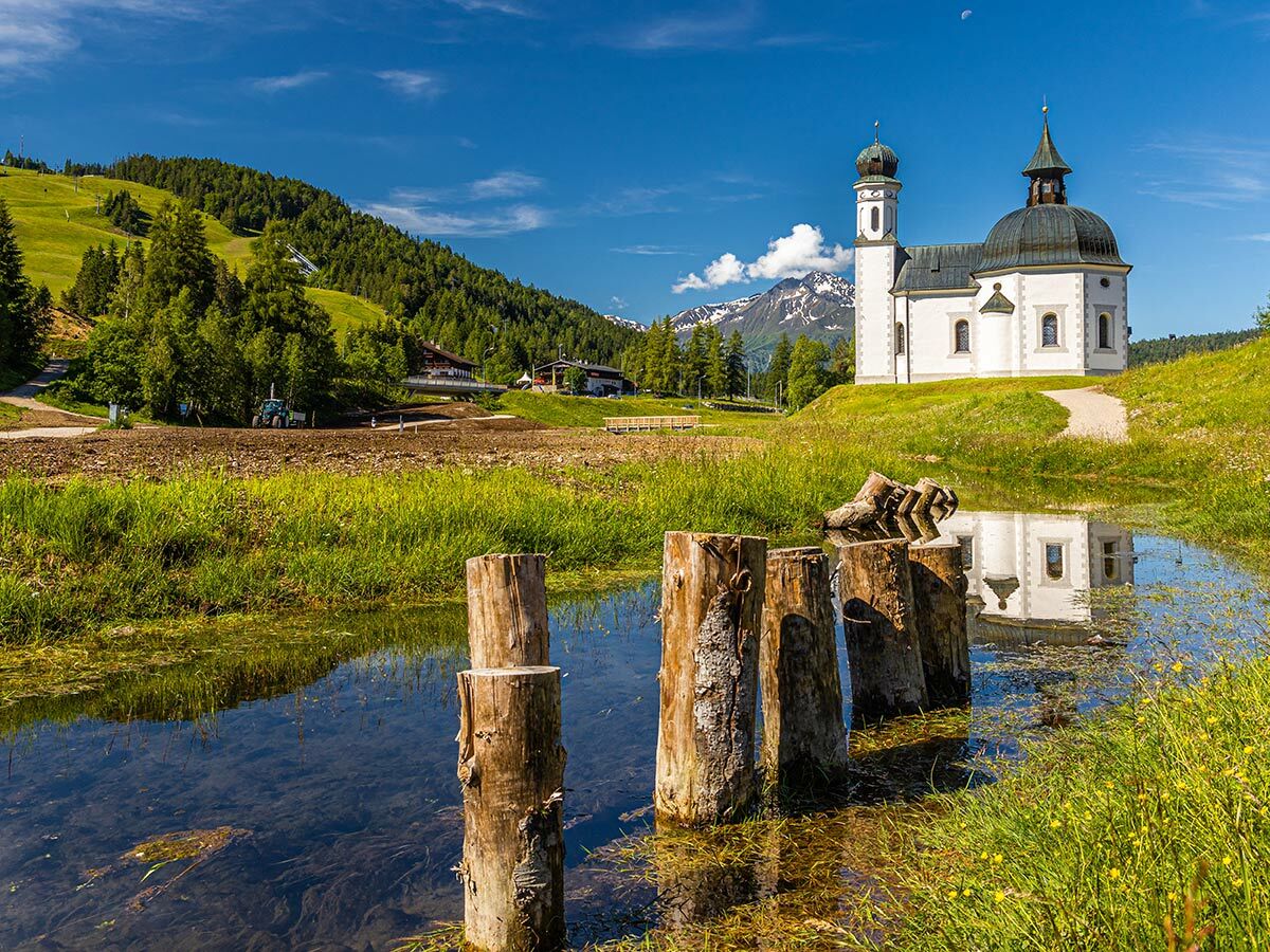 Malerische Alpenkirche am Seeufer mit Holzpfählen, umgeben von grünen Wiesen und schneebedeckten Bergen unter blauem Himmel.
