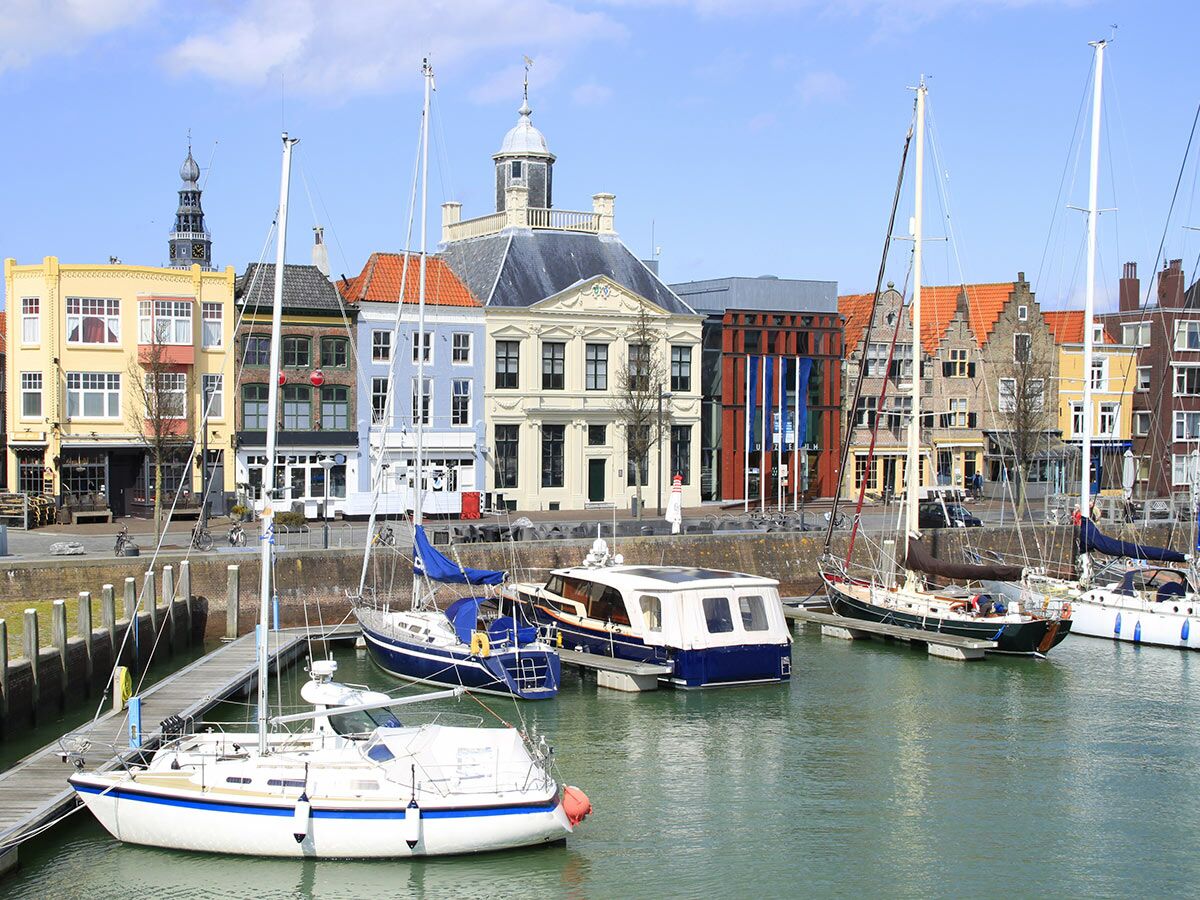 Blick auf einen malerischen Hafen mit Segelbooten und Motorbooten, umgeben von bunten, historischen Gebäuden und einem Rathausturm unter blauem Himmel.