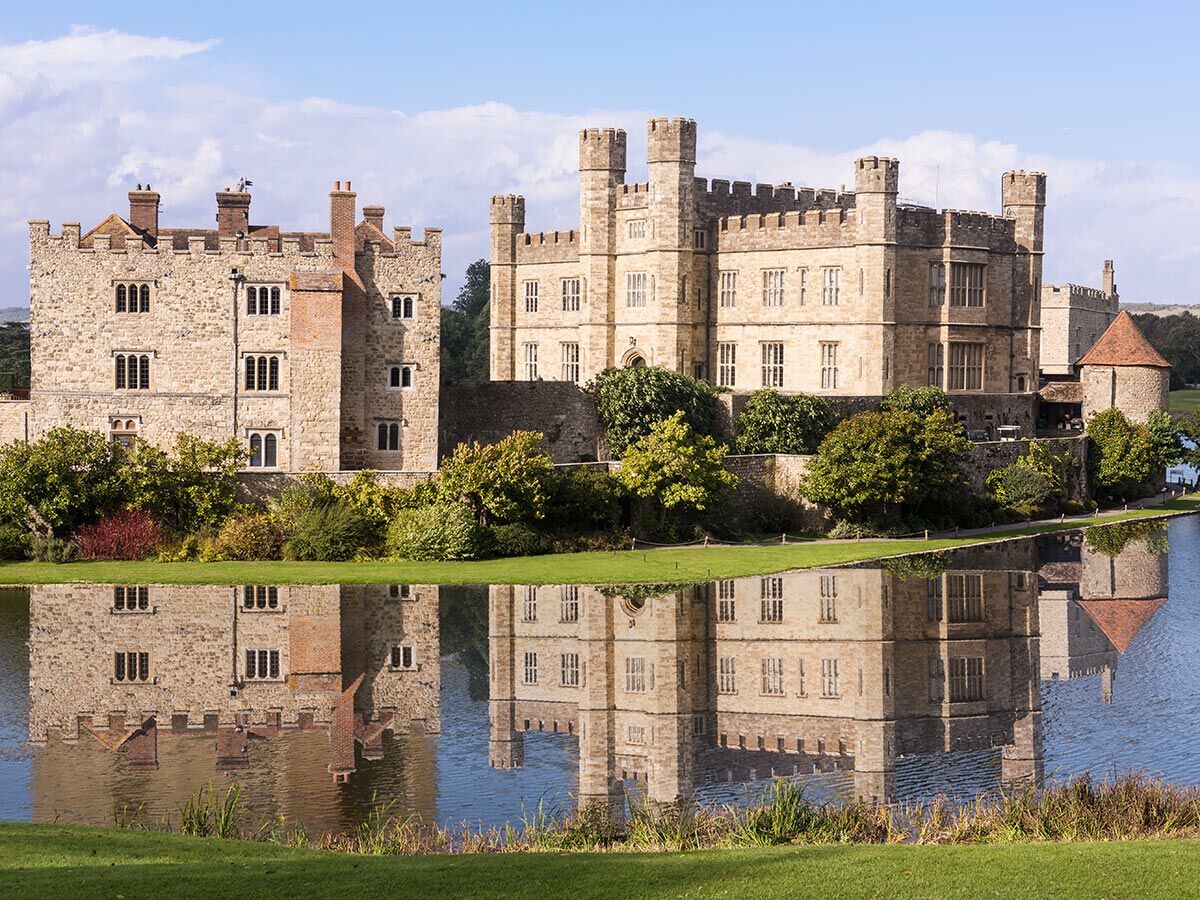 Historisches Leeds Castle aus Stein mit Zinnen, umgeben von Bäumen, perfekt im ruhigen Wasser unter blauem Himmel gespiegelt.
