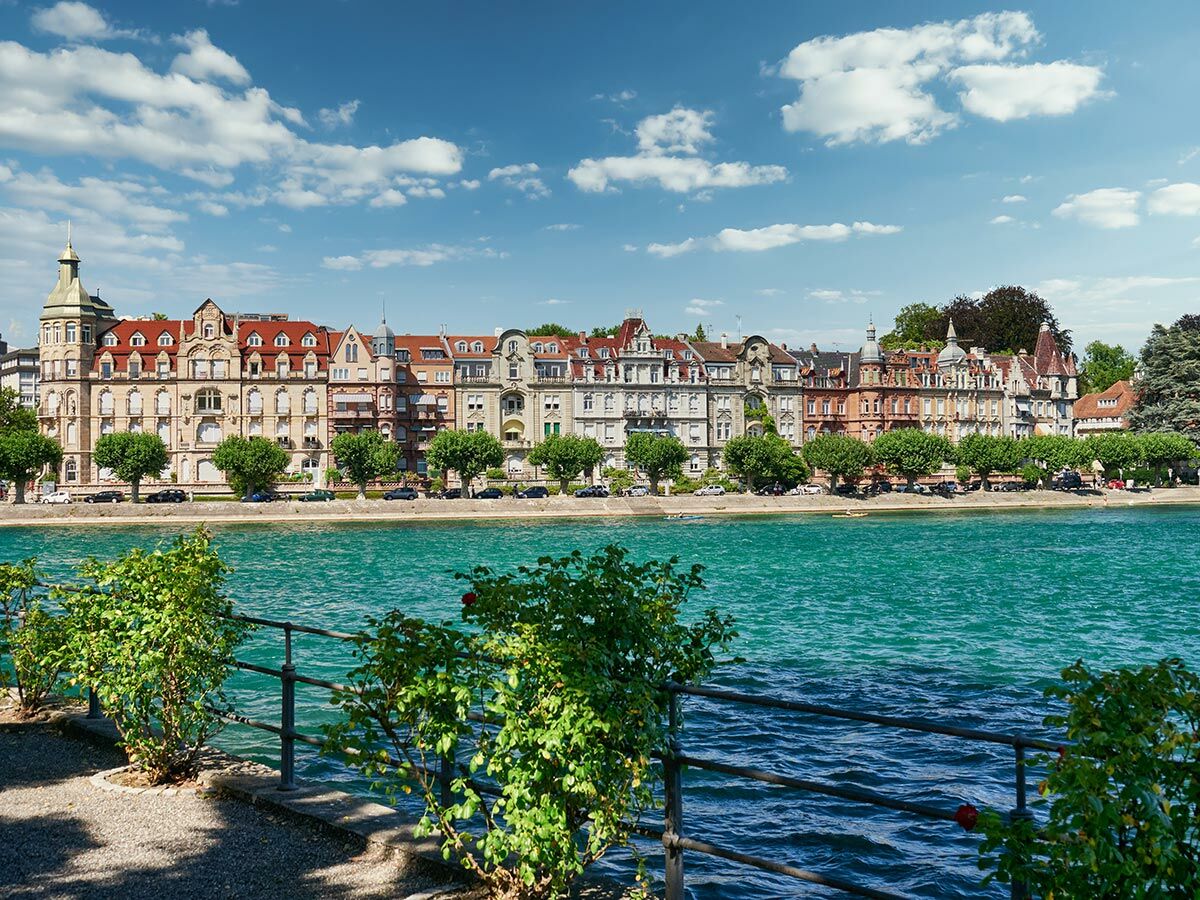 Historische Gebäude und grüne Bäume säumen die Uferpromenade eines Sees mit türkisfarbenem Wasser unter blauem Himmel.