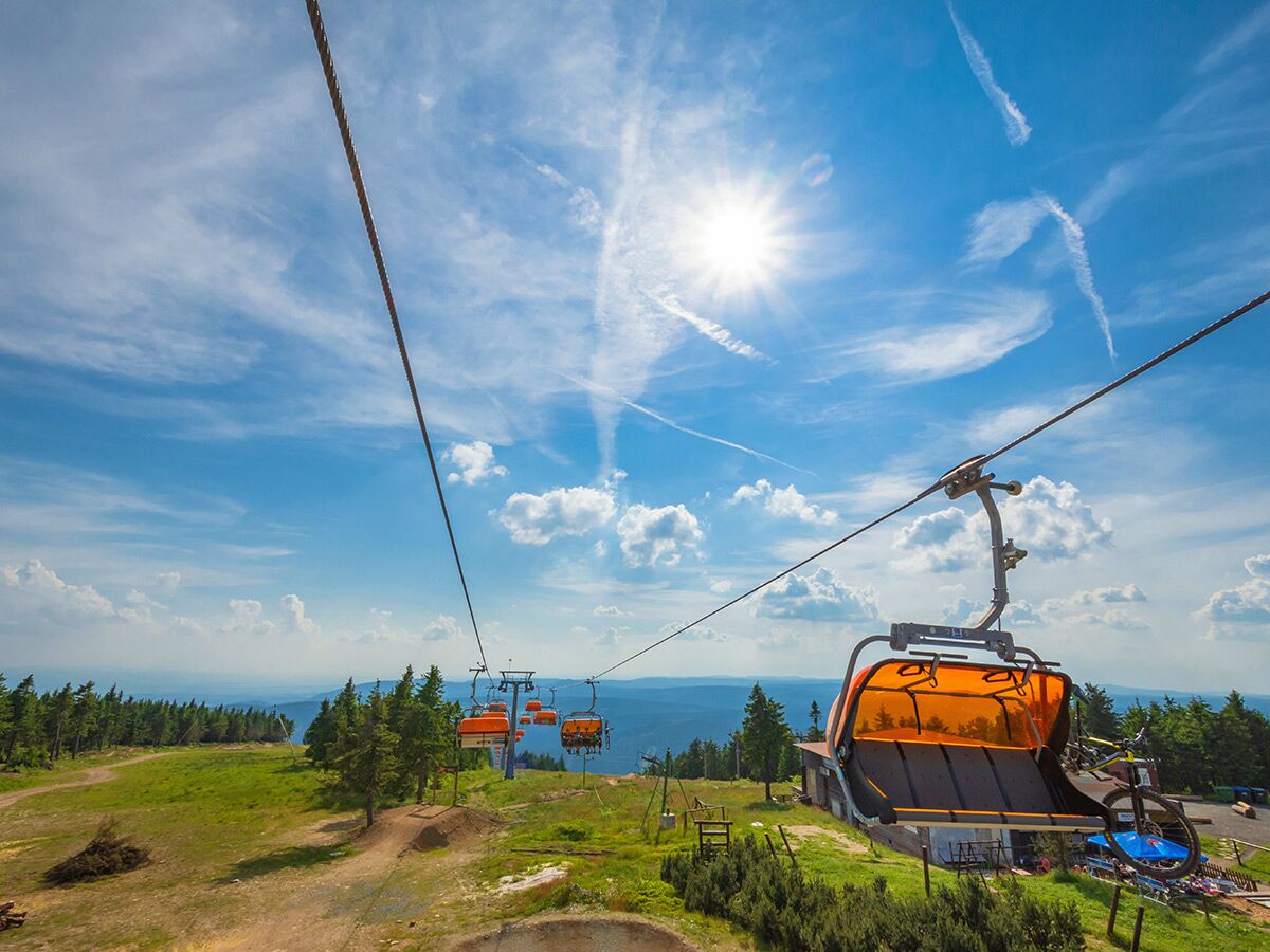 Orange Sessellifte mit Mountainbikes schweben über einer grünen Berglandschaft unter blauem Himmel mit Sonne.