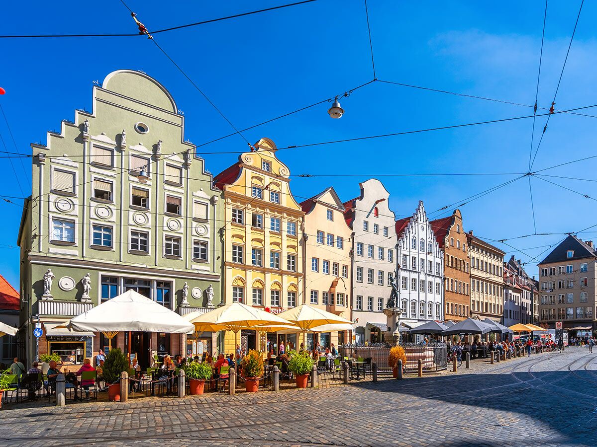 Bunte Giebelhäuser an einem belebten Stadtplatz in Augsburg mit Straßencafés und vielen Menschen unter blauem Himmel.