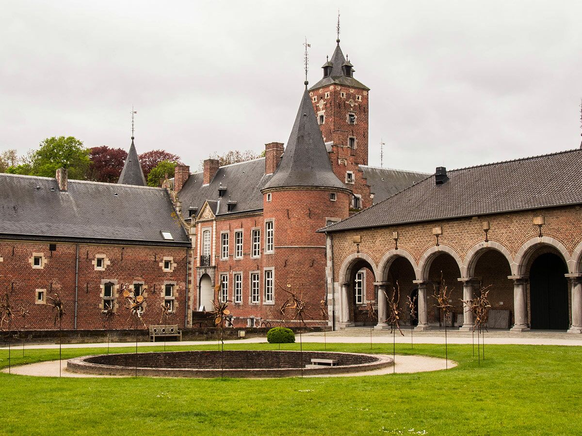 Ansicht eines historischen Backstein-Schlosses mit mehreren Gebäuden, Türmen und einem grünen Innenhof mit modernen Skulpturen.