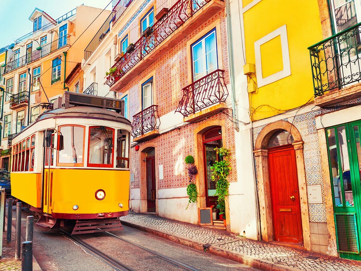 Historische gelbe Straßenbahn (Elétrico 28) fährt durch eine malerische Gasse in Lissabon mit farbenfrohen Gebäuden und Azulejos.