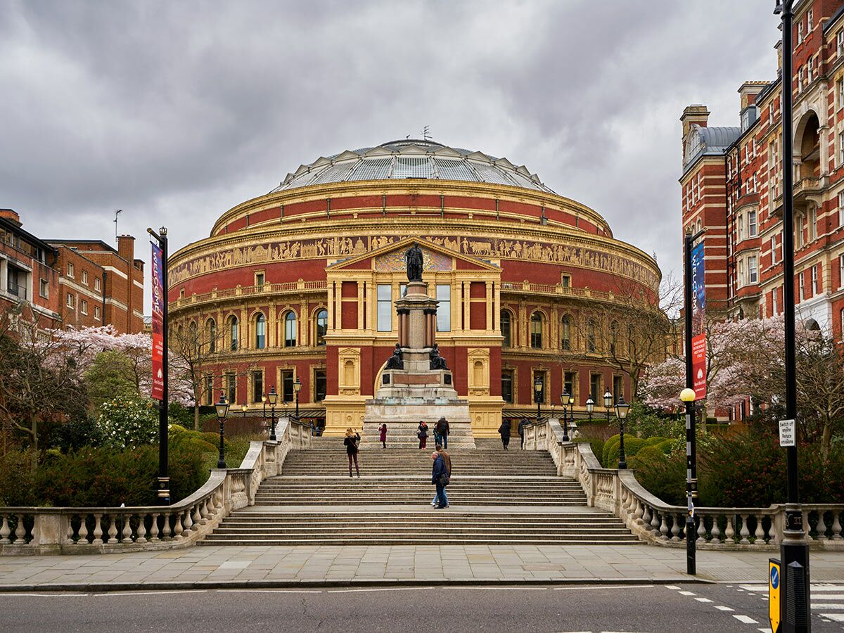 Die Royal Albert Hall in London, ein ikonisches Rundgebäude aus rotem Backstein, umgeben von Frühjahrsgrün und Kirschblüten.