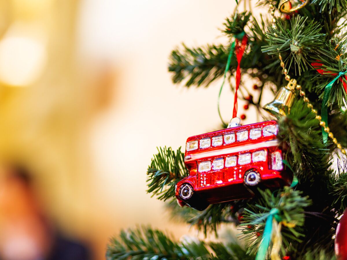 Rotes Londoner Doppeldeckerbus-Ornament hängt an einem grünen Weihnachtsbaum mit goldenen Glocken und Bändern, festliche Deko.