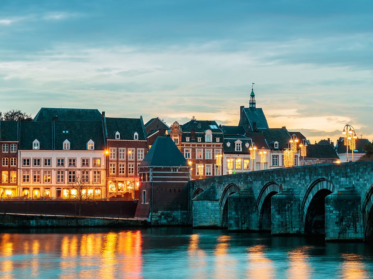 Abendliche Ansicht der Sint Servaasbrücke in Maastricht über die Maas, mit beleuchteten Gebäuden und Reflexionen im Wasser.