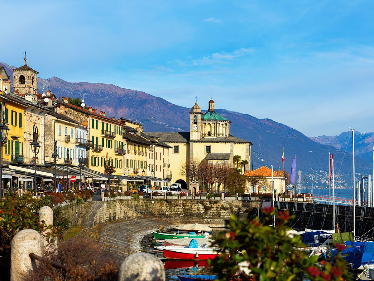 Malerische Hafenstadt Cannobio am Lago Maggiore mit bunten Gebäuden, Kirche, Bergen und Booten unter blauem Himmel.