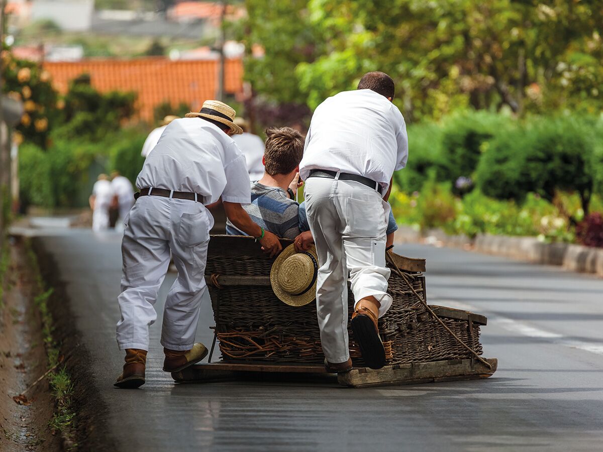 Zwei traditionelle Korbschlittenfahrer in weißen Uniformen schieben einen Touristen in einem Weidenkorbschlitten die Straße hinunter auf Madeira.