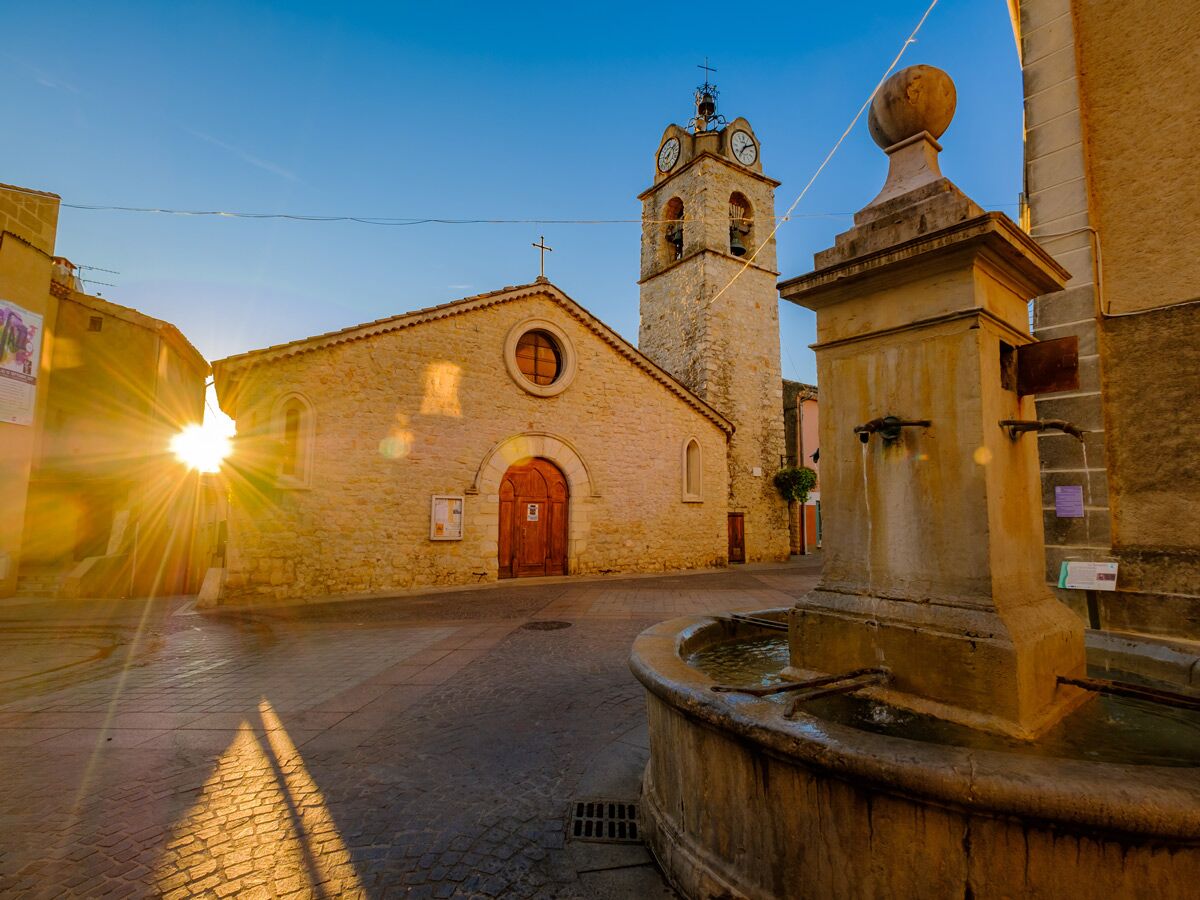 Goldener Sonnenuntergang über einem historischen Dorfplatz mit steinerner Kirche, Glockenturm, Uhr und altem Brunnen in Südeuropa.