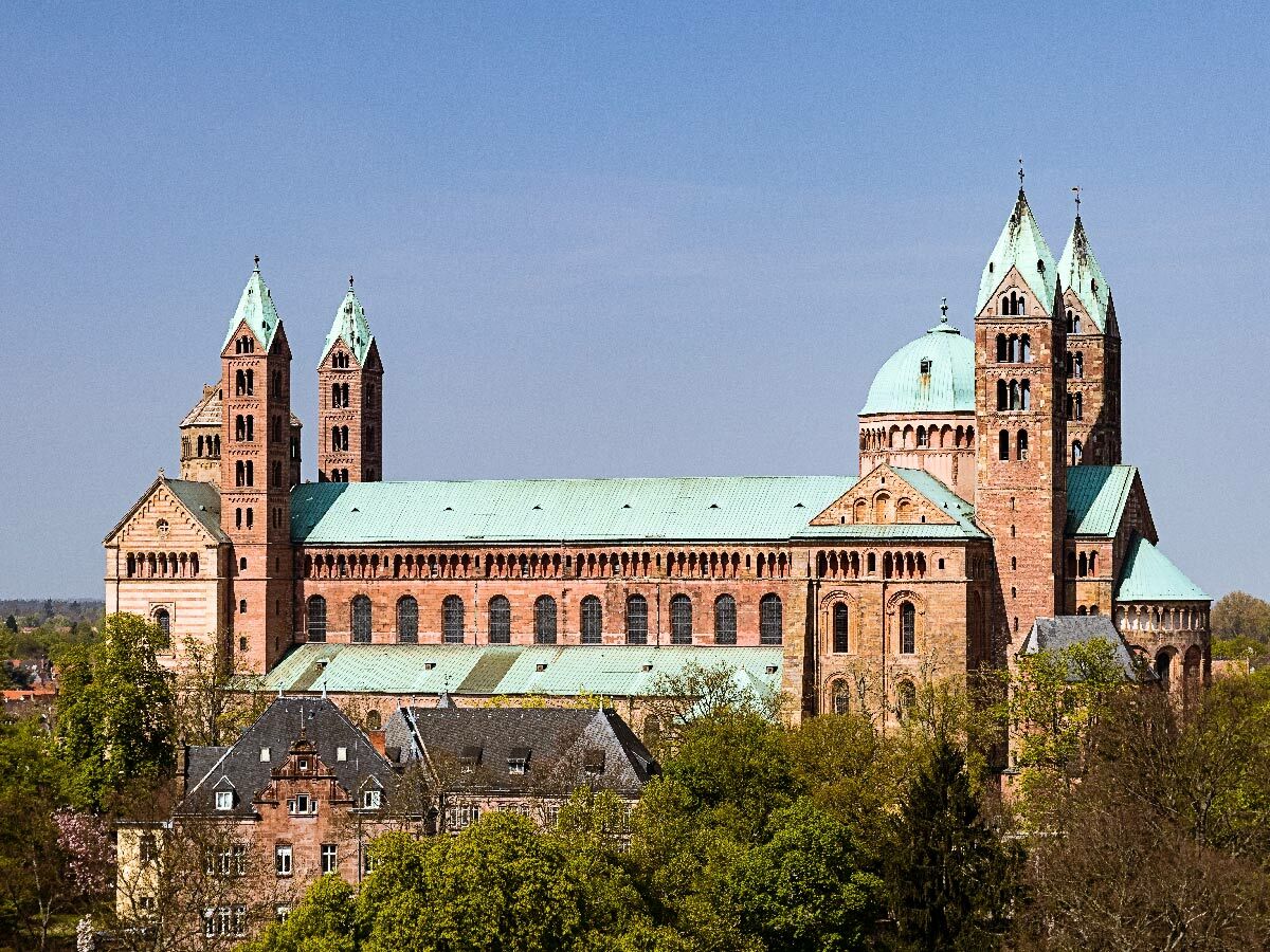 Panoramablick auf den Speyerer Dom, eine beeindruckende romanische Kathedrale mit grünen Kupferdächern und Türmen, umgeben von Bäumen.