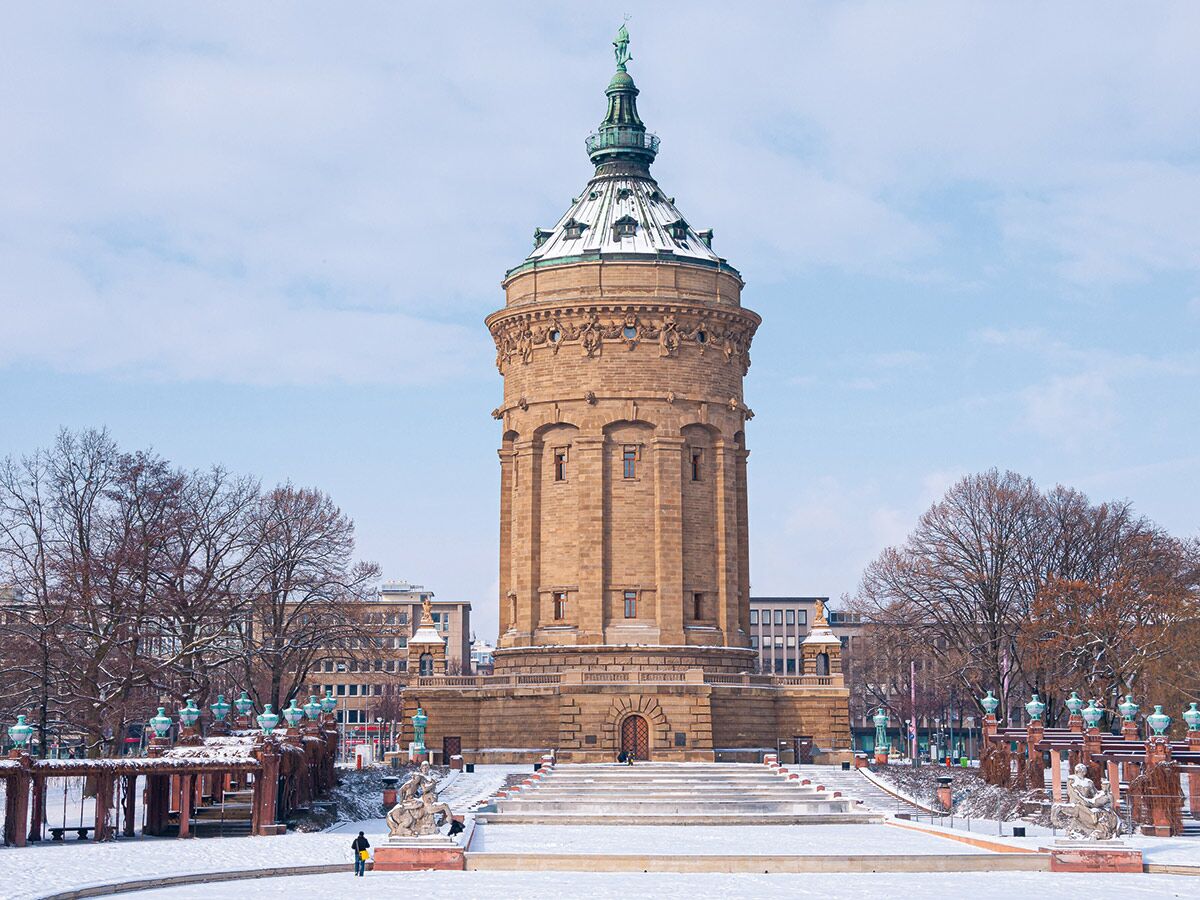 Historischer Wasserturm Mannheim im winterlichen Park mit Schnee, umgeben von Bäumen und barocken Skulpturen unter blauem Himmel.